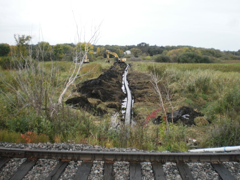 railroad in front, prairie in background with ground dug out, new culvert being placed inside 
